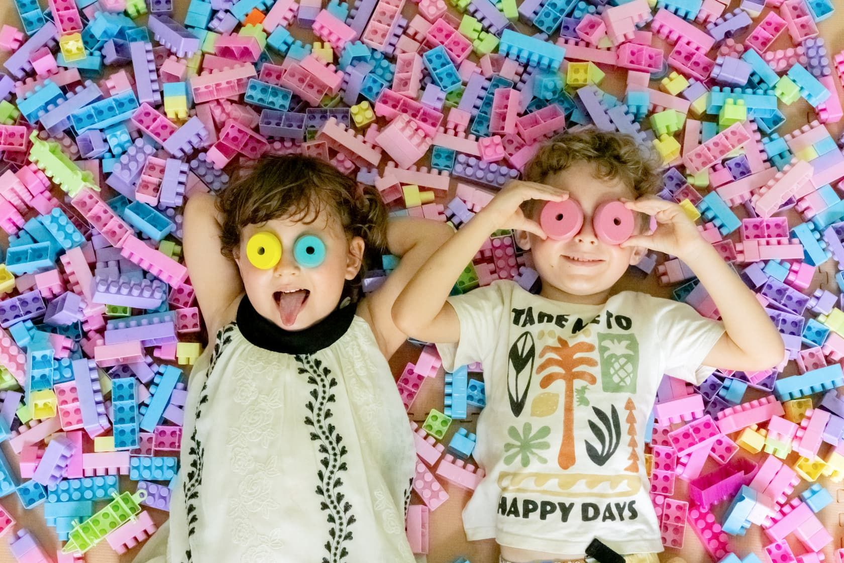Two children lying on a pile of colourful building blocks, holding toy rings over their eyes